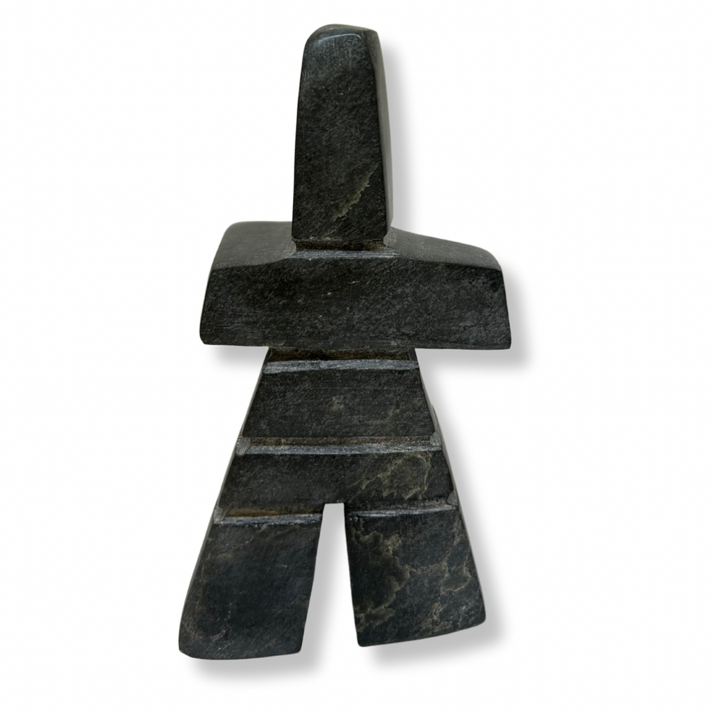 An angular inukshuk in the shape of a person standing in with feet shoulder-width apart. The inukshuk is carved from black stone with stones tightly fitted to each other.