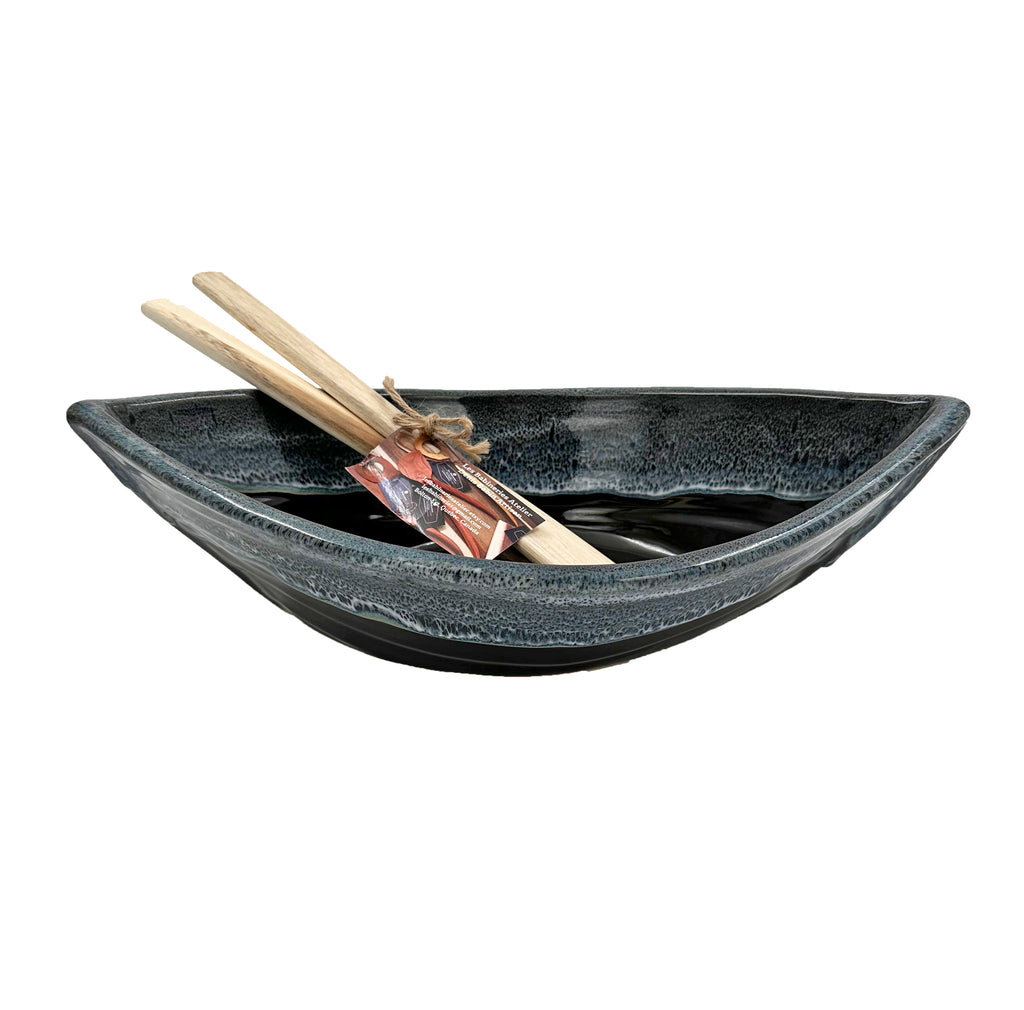Bottom half of bowl is black. Top half of bowl is dark blue with white accents. Bowl is oval shaped with points at two opposite ends. Two wooden paddles in the bowl.