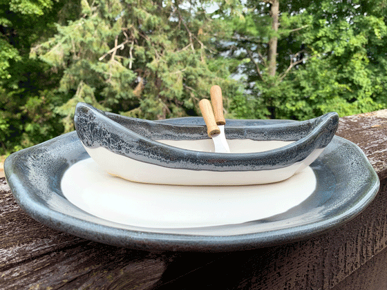 A stoneware canoe sits on a matching stoneware serving plate atop a weathered wooden railing. Two spreaders shaped like paddles rest against the gunnel. The base of both the canoe and platter are off-white, with contrasting rims in mottled blue and white. A peaceful background of trees and lake complete the picture.