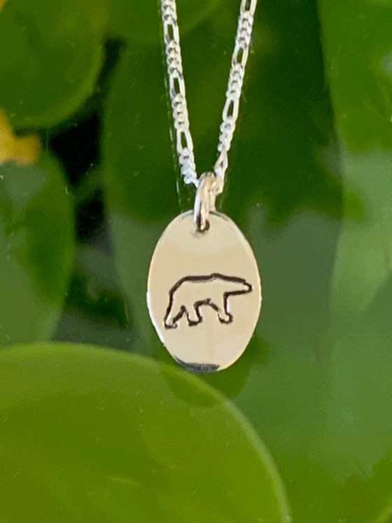 A necklace made of sterling silver hangs freely in front of a forest backdrop. The pendant is an oval of bright polished silver with a polar bear etched into the centre.