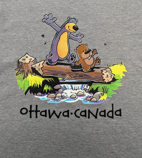 A grey shirt with a dark grey bear with a yellow stomach and a small brown beaver. They are using a piece of wood as a plank to cross a small stream of water. Underneath is written "Ottawa" and "Canada"