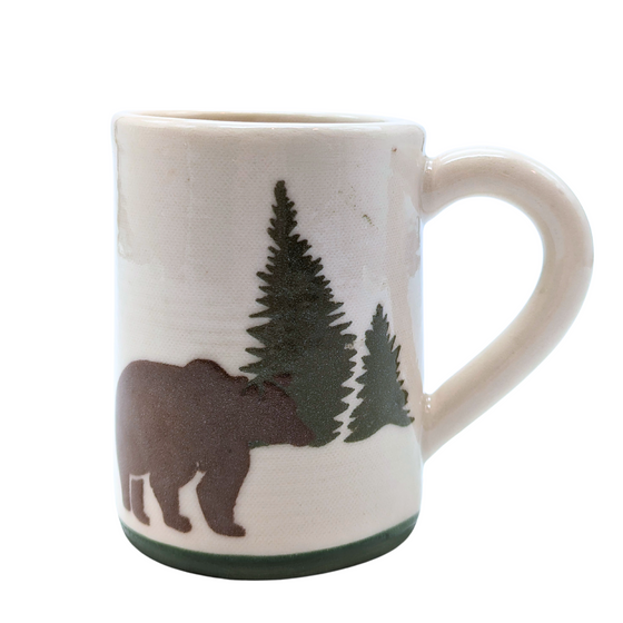 A handformed stoneware mug of white clay with ergonomic handle. The base is green. In the foreground, a brown bear stands in front of two spruce pines of different heights, handpainted with a mixture of traditional brush and sgraffito on the side.
