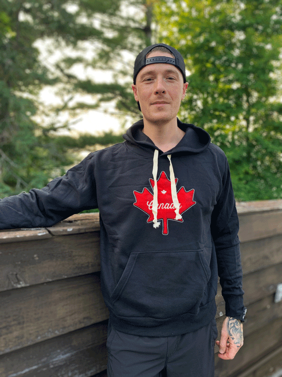 A black hoodie with white drawstrings. A red maple leaf is on the chest, with Canada and a smaller maple leaf written in white across it.