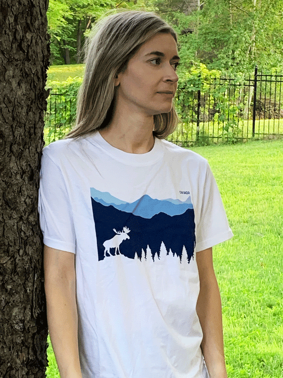 A white shirt. In the middle are mountains of different shades of blue. There is a white moose and trees at the bottom of the mountains. In the top right corner is text saying "Canada".