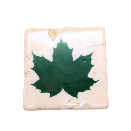 A White stone square coaster. There is a green maple leaf in the middle of the coaster, and there is a shiny coat around it.
