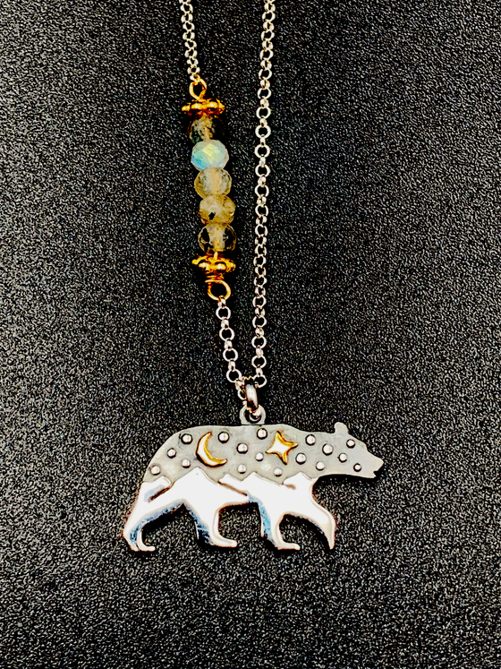 Silver chain with 5 coloured beads in middle of chain attached with gold chain links that look like flowers. Pendant is a bear with legs that turn into mountain peaks. The upper body of the bear resembles a night sky with a  gold cresent moon and a gold 4-point star. Small silver dots are also on the upper body of the bear.