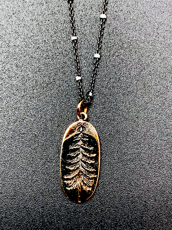 Black chain with thin hexagonal silver rings that are evenly distributed throughout the chain. Pendant is a gold-bronze oval with the indentation of a pine tree. The indentation is black.
