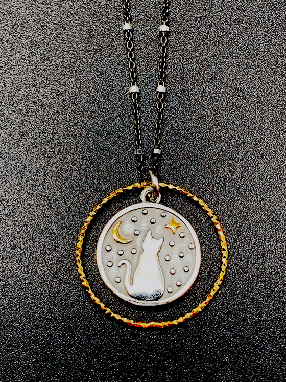Black chain with thin hexagonal silver rings that are evenly distributed throughout the chain. Pendent is a gold ring that has triangle ridges. A smaller circle with a silver outline and a grey background is inside the ring. There is a cat sitting and facing away in the center of the smaller circle. The cat is surrounded by small silver dots and a gold crescent moon and 4-point star.  