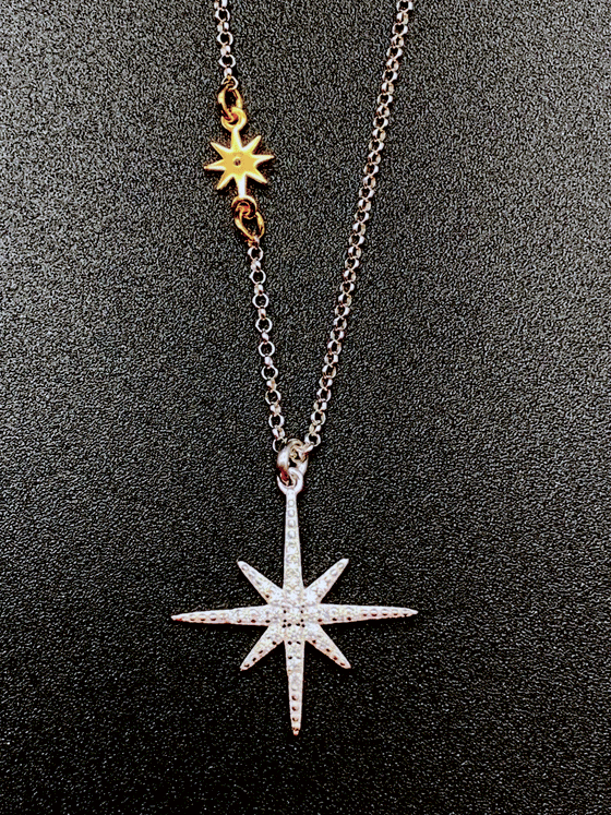 Silver chain with small gold star with eight even points attached in the middle of the of the chain. Gold star is symmetrical. Larger star is the pendent with jewels covering the top surface. Silver star also has 8 points. The points that point up, down, left, and right are longer. Necklace is on a black background.