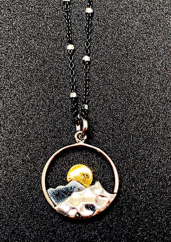 Black chain with thin hexagonal silver rings that are evenly distributed throughout the chain. Pendant is a silver outline of a circle. At the bottom of the outline is a silver mountain attached to the outline. The mountain has grooves to make it realistic. Behind the silver mountain is a small, flat, grey mountain with a yellow sun showing behind the peak of the mountain. 