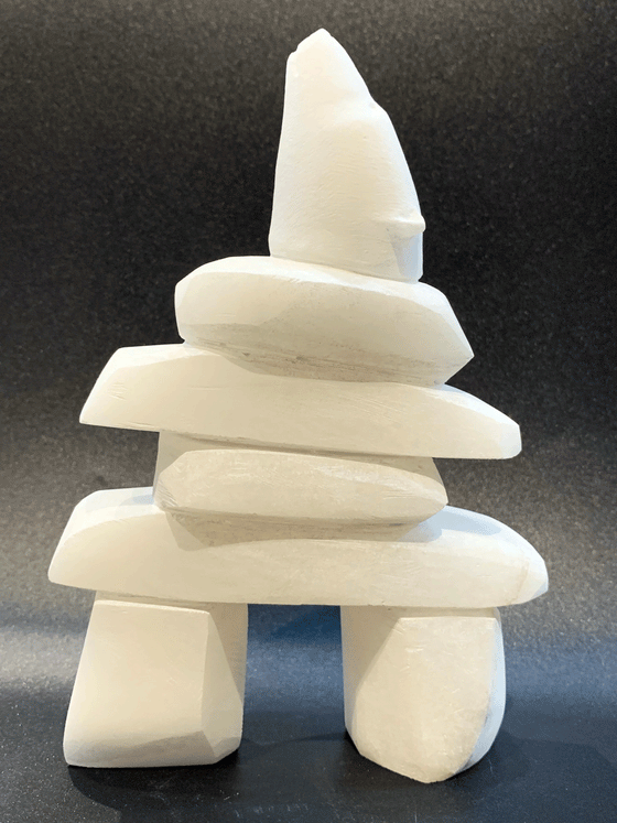 An inukshuk, the top stone of which is a polar bear head looking up, carved in one piece from white alabaster. The piece faces left in this image. The artist has used a special technique to etch fur texture into the bear and carving marks into the stone of the inukshuk for added realism.
