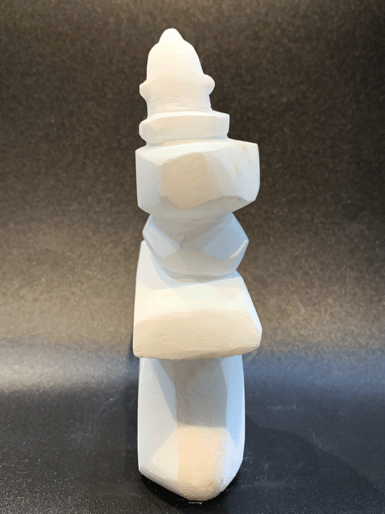 An inukshuk, the top stone of which is a polar bear head looking up, carved in one piece from white alabaster. The piece faces away in this image. The artist has used a special technique to etch fur texture into the bear and carving marks into the stone of the inukshuk for added realism.