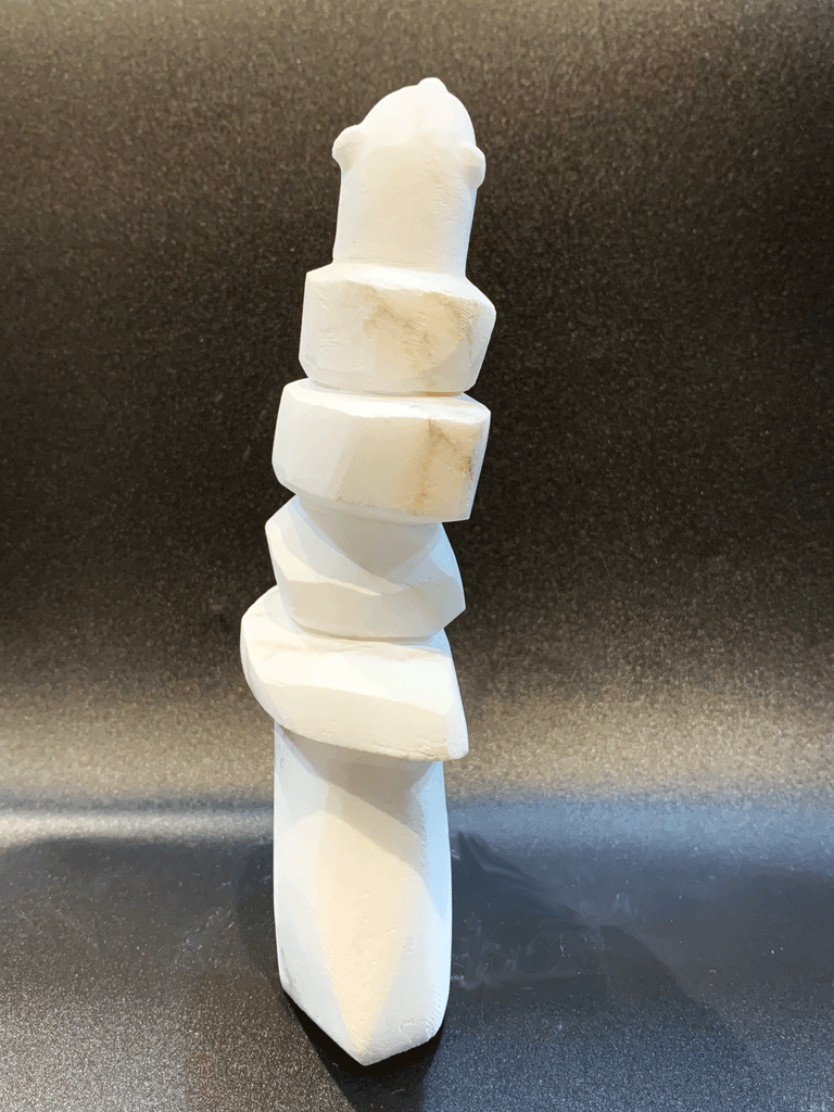 An inukshuk, the top stone of which is a polar bear head looking up, carved in one piece from white alabaster. The piece faces away in this image. The artist has used a special technique to etch fur texture into the bear and carving marks into the stone of the inukshuk for added realism.