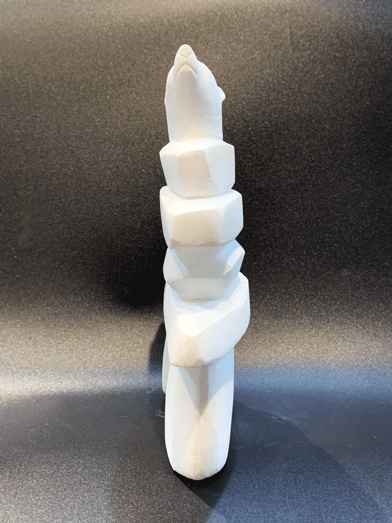 An inukshuk, the top stone of which is a polar bear head looking up, carved in one piece from white alabaster. The piece faces straight ahead in this image. The artist has used a special technique to etch fur texture into the bear and carving marks into the stone of the inukshuk for added realism.