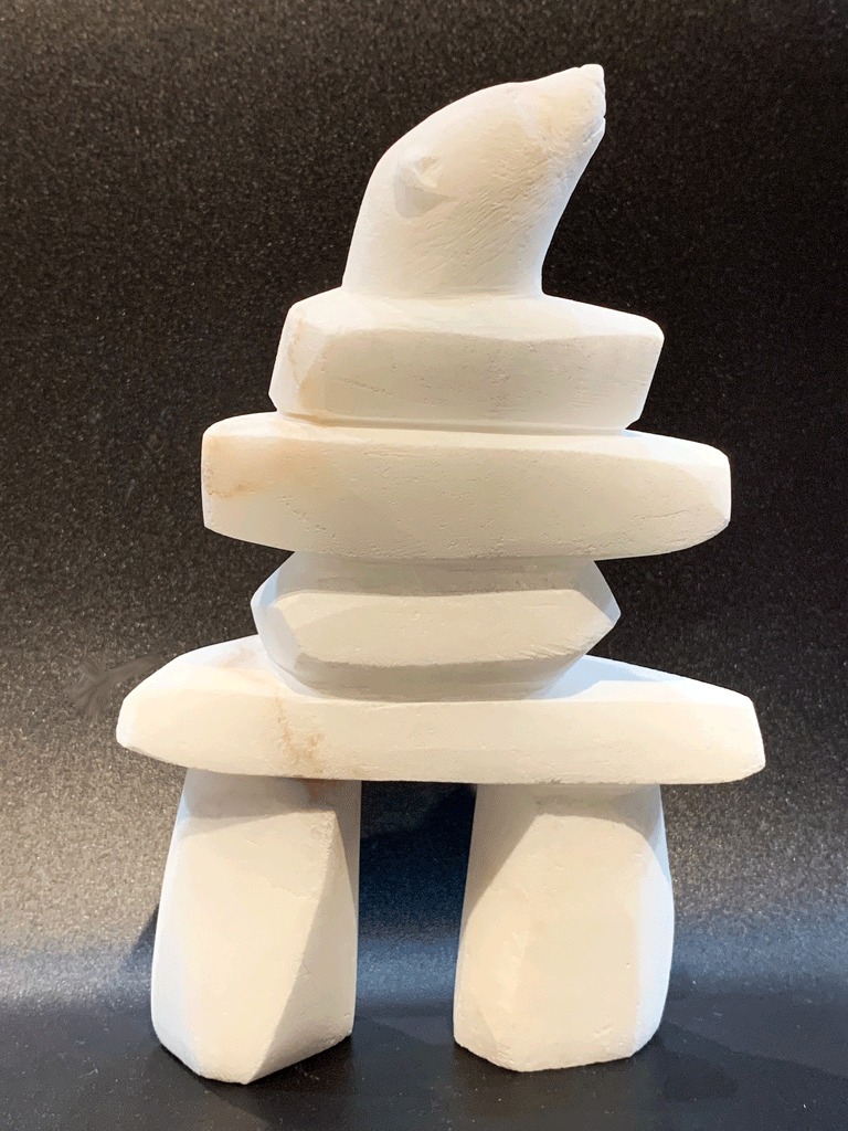 An inukshuk, the top stone of which is a polar bear head looking up, carved in one piece from white alabaster. The piece faces right in this image. The artist has used a special technique to etch fur texture into the bear and carving marks into the stone of the inukshuk for added realism.