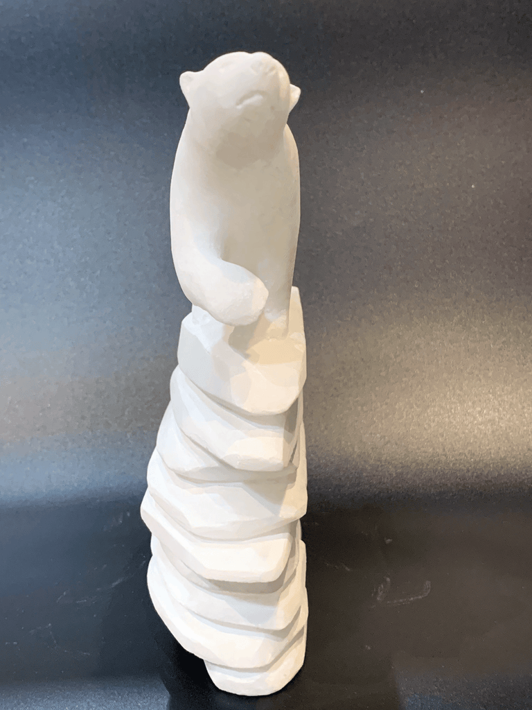 A polar bear standing on top of an inukshuk base, carved in one piece from white alabaster. The polar bear faces straight ahead with one forepaw raised. The artist has used a special technique to etch fur texture into the bear and carving marks into the stone of the inukshuk for added realism.