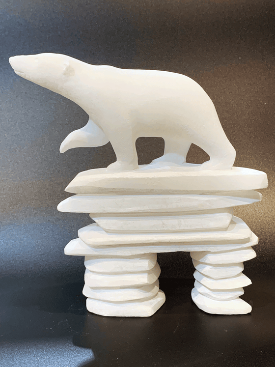 A polar bear standing on top of an inukshuk base, carved in one piece from white alabaster. The polar bear faces left with one forepaw raised. The artist has used a special technique to etch fur texture into the bear and carving marks into the stone of the inukshuk for added realism.