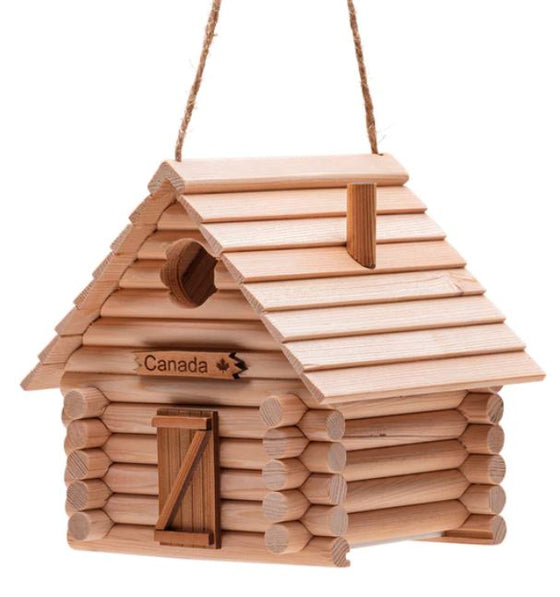 A birdhouse made of interlocking logs, featuring a traditional Z-panel door, a chimney, and a sign reading "Canada" is mounted above the door. The space above the sign has a small hole for songbirds.