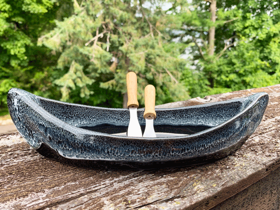 A stoneware canoe sits on a weathered wooden railing. Two spreaders shaped like paddles rest against the gunnel. The lower half of the canoe is black, with a contrasting rim in mottled blue, black, and white. A peaceful background of trees and lake complete the picture.