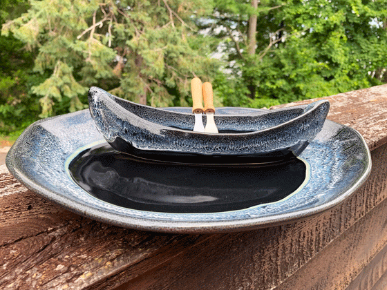 A stoneware canoe sits on a matching stoneware serving plate atop a weathered wooden railing. Two spreaders shaped like paddles rest against the gunnel. The base of both the canoe and platter are black, with contrasting rims in mottled blue and white. A peaceful background of trees and lake complete the picture.