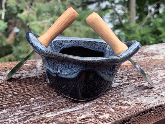 A stoneware rowboat sits on a weathered wooden railing. Two spreaders shaped like paddles sit in the oarlocks. The base is black, with a contrasting rim in mottled blue, black, and white. A peaceful background of trees and lake complete the picture.