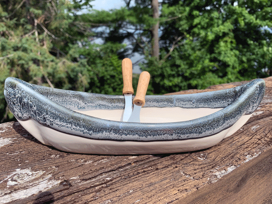 A stoneware canoe sits on a weathered wooden railing. Two spreaders shaped like paddles rest against the gunnel. The lower half of the canoe is off-white, with a contrasting rim in mottled blue and white. A peaceful background of trees and lake complete the  picture.