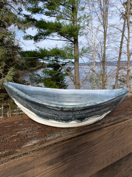 Bottom half of bowl is off-white. Top half of bowl is blue with white accents. Bowl is oval shaped with points at two opposite ends. Bowl is on a piece of wood with trees and a lake in the background.