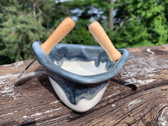 A stoneware rowboat sits on a weathered wooden railing. Two spreaders shaped like paddles sit in the oarlocks. The base is white, with a contrasting rim in mottled blue, black and white. A peaceful background of trees and lake complete the picture.