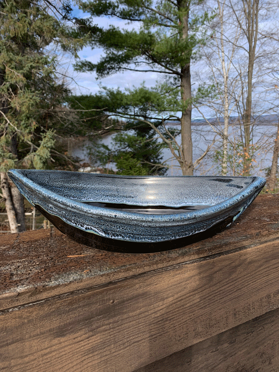 Bottom half of bowl is black. Top half of bowl is dark blue with white accents. Bowl is oval shaped with points at two opposite ends. Bowl is on a piece of wood with trees and a lake in the background.