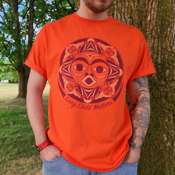 An orange shirt. In the middle there is an indigenous design resembling a face. The design is in the shape of a circle and underneath is written "Every Child Matters" in cursive font. The design and text is in purple and there are light orange details.