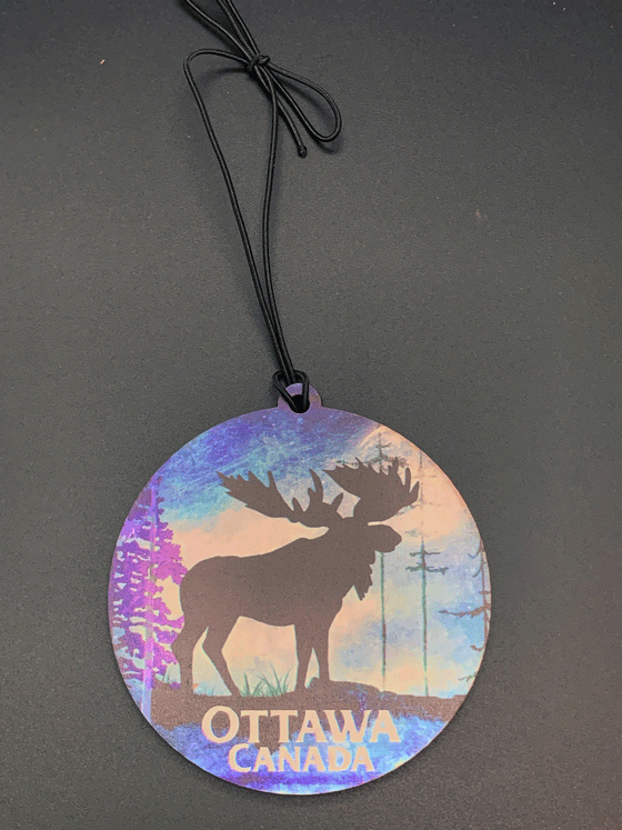 A flat, circular wooden ornament featuring a black moose standing against a blue, purple and white forest background. The words "Ottawa, Canada" are written in white below the moose.
