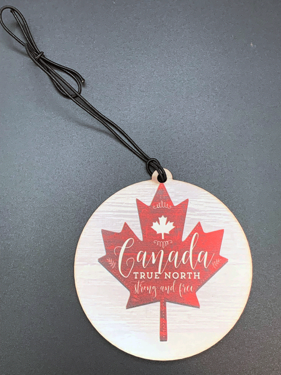 A flat, circular wooden ornament with a red maple leaf against a white background. White text written on the maple leaf reads "Canada True North Strong and Free".