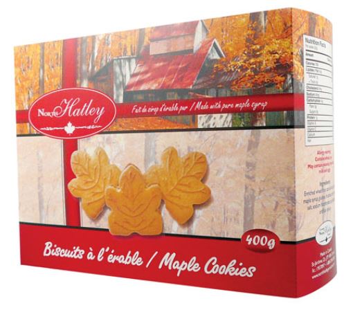 The box is printed to mimic a red ribbon wrapping, dividing the box into quadrants depicting autumn scenes of a sugar shack. In the foreground, three leaf-shaped maple cream cookies draw the eye.