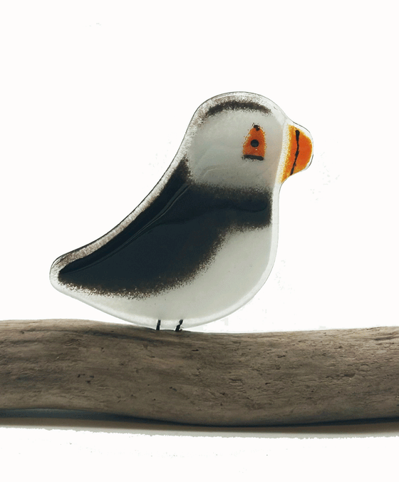 2-D black and white Puffin. Has orange beak and eye. Has 2 small metal rods coming from the bottom of it that look like legs. The metal rods are attached to a piece of a tree that looks like a small tree stump. The background is white.