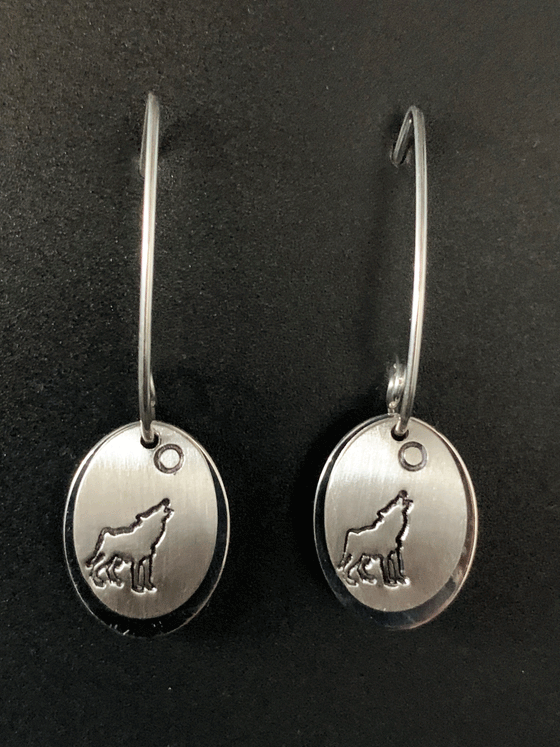 Silver earrings made of two layers of brushed and polished silver mounted on sterling silver hooks. The base is an oval of bright polished silver. Over that is a smaller circle of brushed silver with a wolf howling at the moon etched into the centre. The posts hook through the two layers and spiral at the end to keep them in place. The hooks feature long backs that are both functional and decorative.