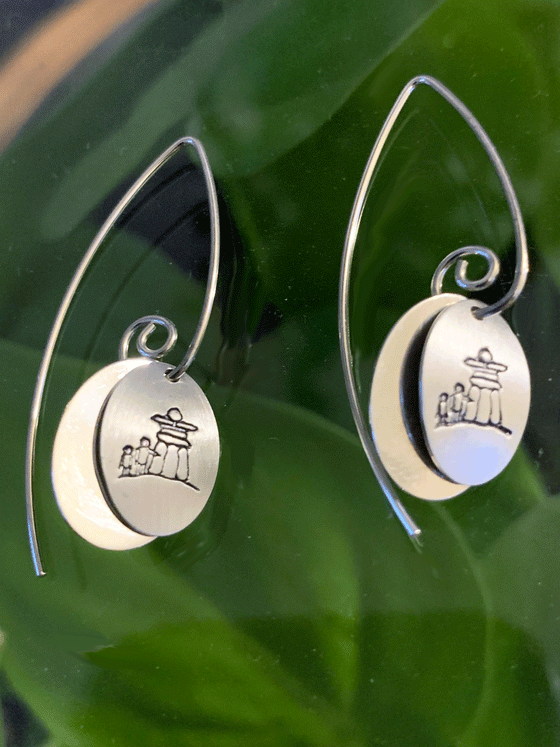 Silver earrings made of two layers of brushed and polished silver mounted on sterling silver hooks. The base is an oval of bright polished silver. Over that is a smaller circle of brushed silver with a large inukshuk and two smaller human figures etched into the centre. The posts hook through the two layers and spiral at the end to keep them in place. The hooks feature long backs that are both functional and decorative.