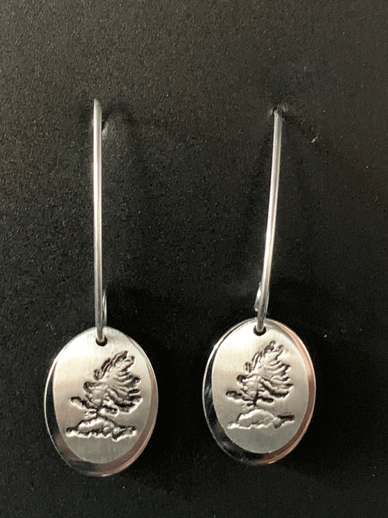 Silver earrings made of two layers of brushed and polished silver mounted on sterling silver hooks. The base is an oval of bright polished silver. Over that is a smaller circle of brushed silver with a rugged windswept pine on rock etched into the centre. The posts hook through the two layers and spiral at the end to keep them in place. The hooks feature long backs that are both functional and decorative.