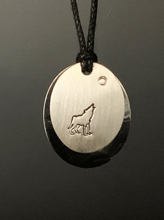 A necklace made of two layers of brushed and polished silver mounted on a leather cord. The base is an oval of bright polished silver. Over that is a smaller circle of brushed silver with a wolf howling at the moon  etched into the centre.