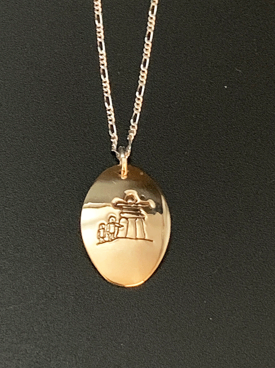 A necklace made of 14 karat gold fill and sterling silver on a gold chain. The base is an oval of bright gold with a large inukshuk with two smaller human figures etched into the centre.