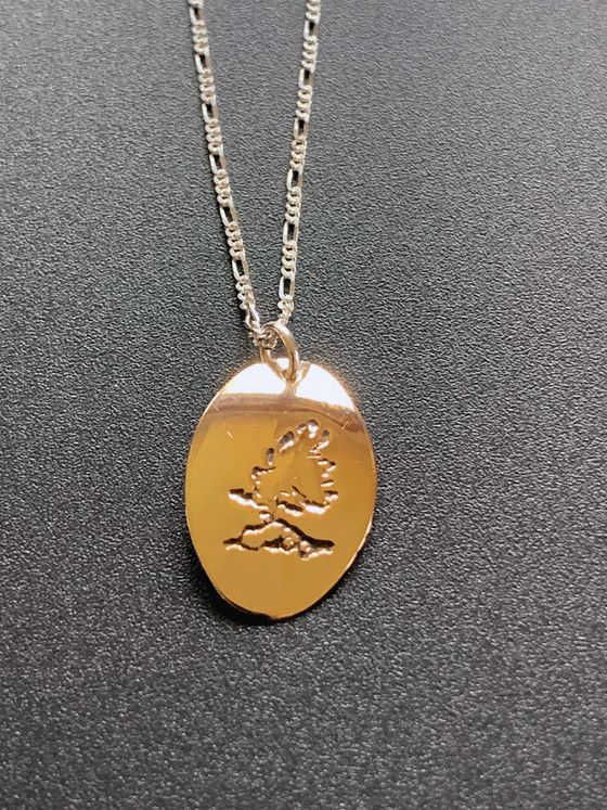 A necklace made of 14 karat gold fill and sterling silver on a gold chain. The base is an oval of bright gold with a rugged windswept pine on rock etched into the centre.