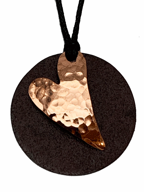 A circular heart necklace mounted on leather cord. The pendant is made of two layers: antique upcycled tin cut into a circle forms the base, and then a hammered copper heart is overlaid to create stunning contrast.