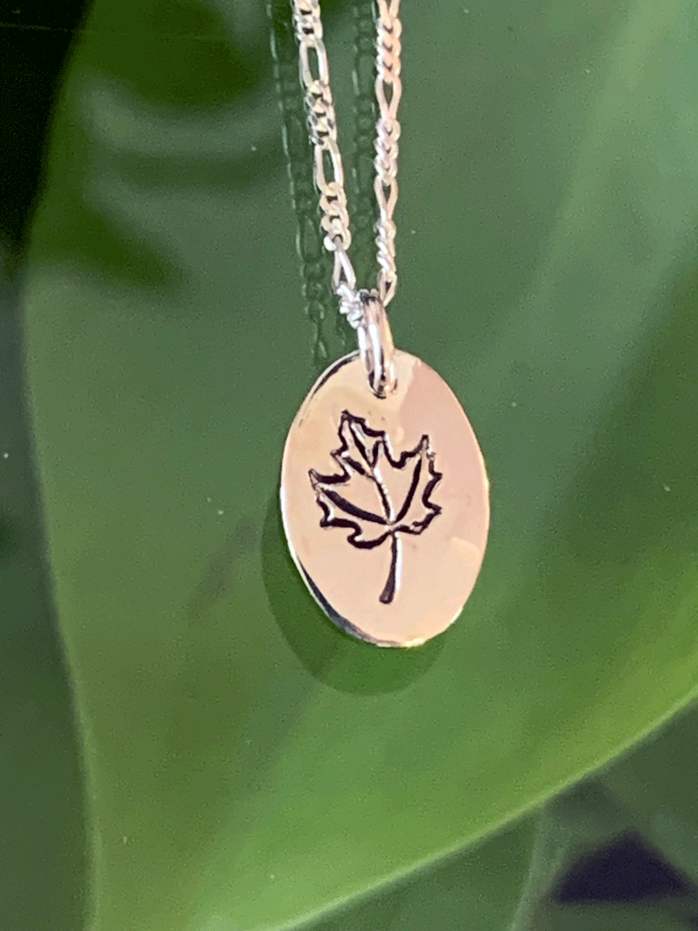 A necklace made of sterling silver hangs freely in front of a forest backdrop. The pendant is an oval of bright polished silver with maple leaf etched into the centre.