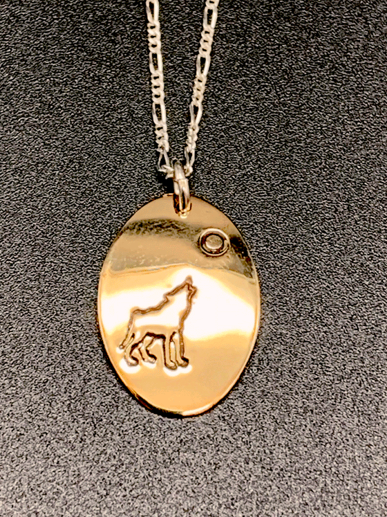 A necklace made of 14 karat gold fill and sterling silver on a gold chain. The base is an oval of bright gold with a wolf howling at the moon etched into the centre.