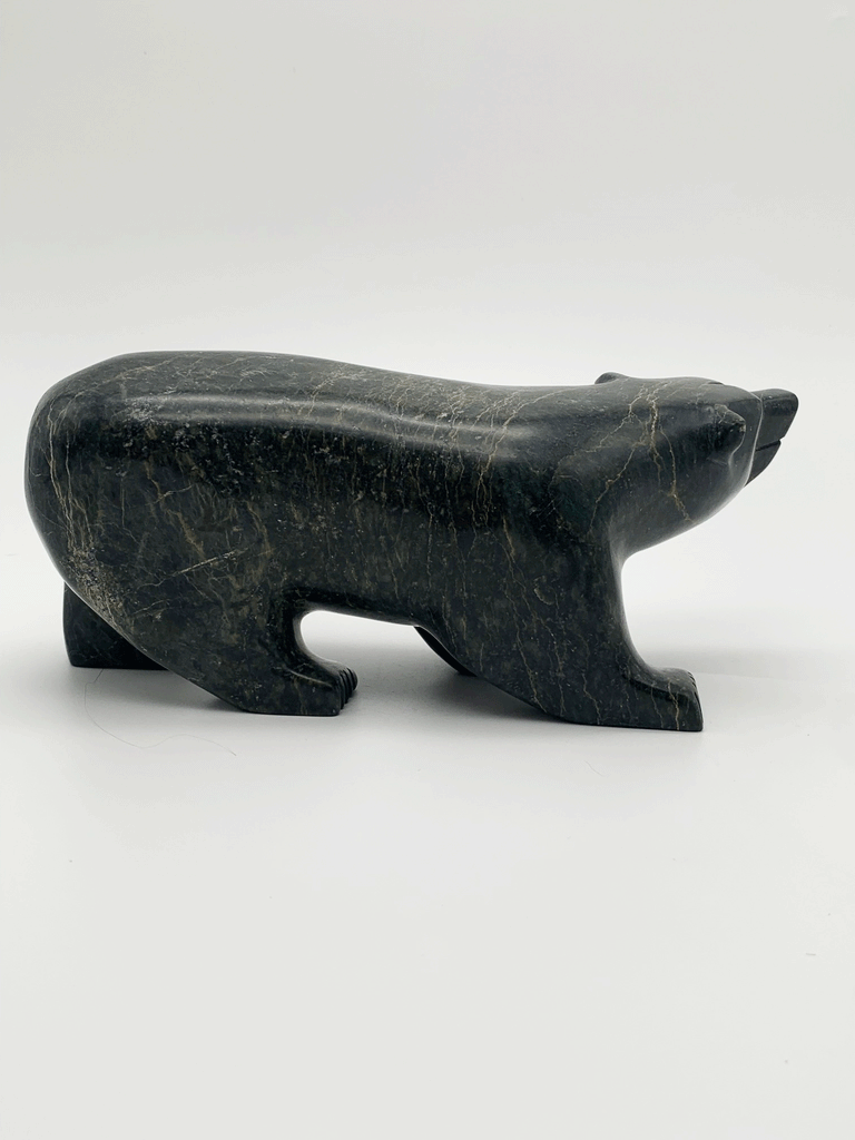 A smiling walking bear carved from very dark green soapstone stands on all fours. Its head is cocked playfully to one side. This bear faces right.