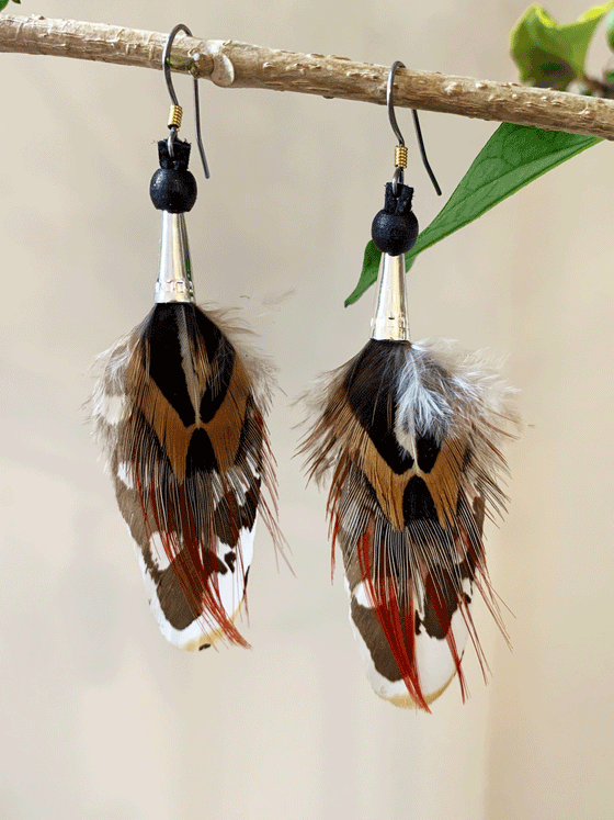 Dangling earrings featuring a black and brown feather with red streaks and white spots, with a silver-coloured stainless steel cone-shaped base and steel hooks.