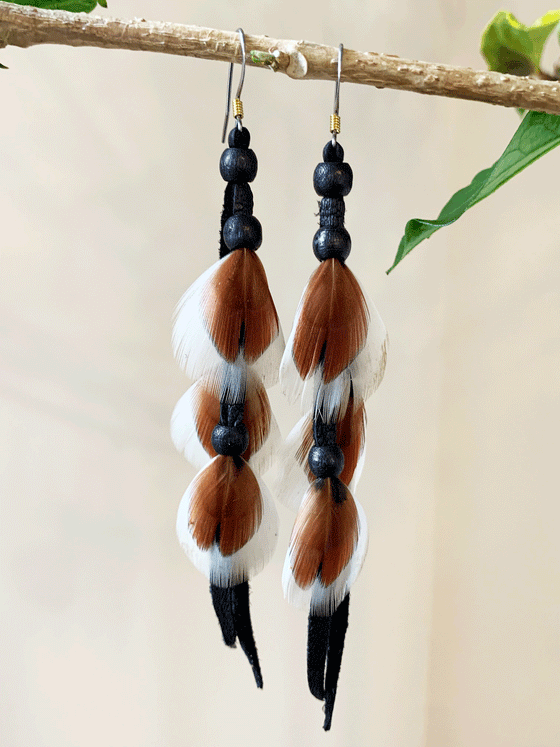 A pair of dangling earrings, each featuring 3 brown and white feathers attached to a black rope with 3 black beads, hanging on a silver-coloured stainless steel hook.