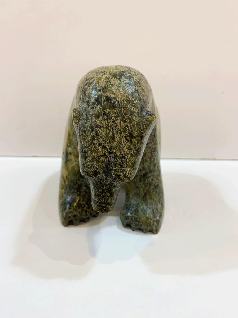 A green and brown soapstone bear stands on all fours, staring ponderously at the ground in front of it. By emphasizing the mass of the head and neck, the artist conveys a sense of power and physical majesty in the animal. This bear faces the viewer straight on.