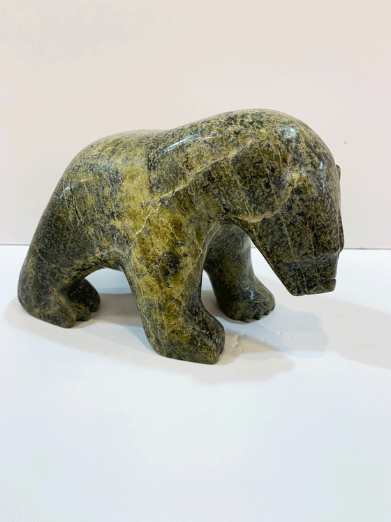 A green and brown soapstone bear stands on all fours, staring ponderously at the ground in front of it. By emphasizing the mass of the head and neck, the artist conveys a sense of power and physical majesty in the animal. This bear faces right.