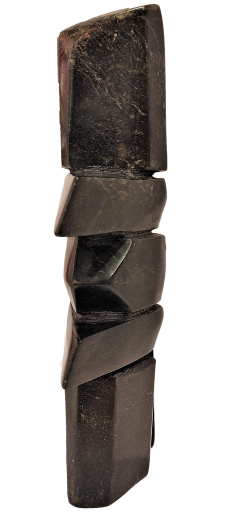 An inukshuk carved from black and brown stone. The legs and head are very tall, while the body is composed of wide slabs forming shoulders and hips, and a rounded stone in the middle.