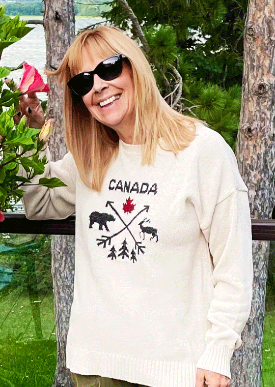 A white crewneck sweater, featuring a black design on the chest: two intersecting arrows with the black silhouette of a deer on the left, three trees on the bottom, a bear on the right, and a red maple leaf on top, and Canada written in black above.